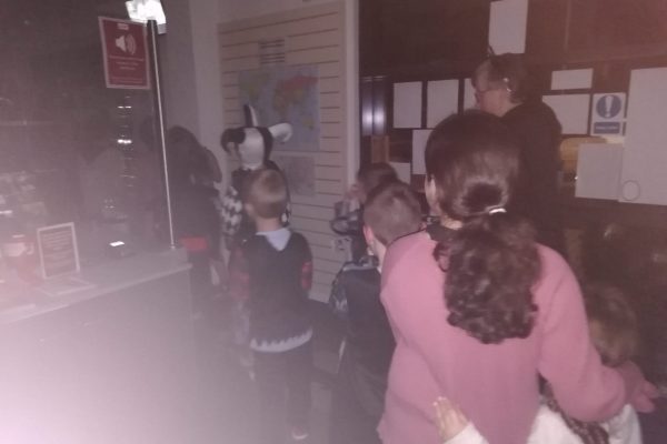 Photo taken in the dark inside the museum, near the entrance. Children in a jumbled assortment of Halloween costumes walk past the front desk in the line.