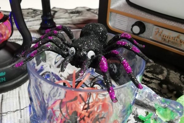 A photo of a glass containing a mystery number of plastic spiders. On top of the glass is a toy spider with googly eyes. In the background part of a Halloween lantern and a toy Halloween TV decoration.