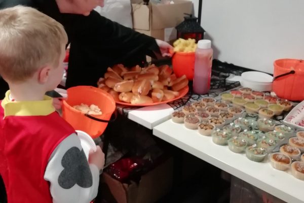 A child dressed as a Dalmatian stands in front of a table containing Halloween themed food in this photo. This includes cupcakes with spiderweb and scary face designs. There is also some hot dogs on the table.