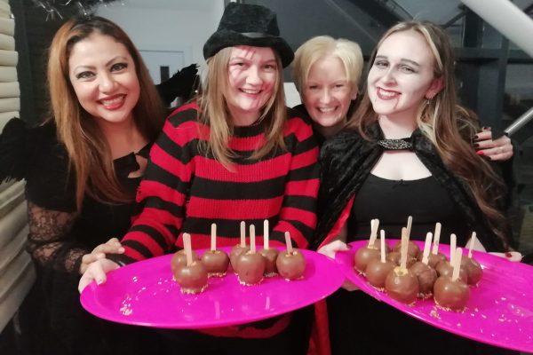 Four happy members of our museum team wearing Halloween costumes. Two of them, dressed as Freddy Krueger and a vampire, hold pink plates full of chocolate apples.