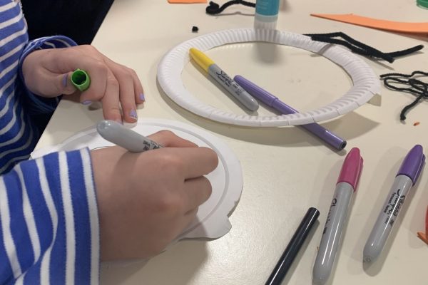 The hands of a young person drawing with felt tip pens on a paper plate. The table they are resting on is littered with craft supplies.