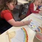 A young person painting a fan with lots of colours.
